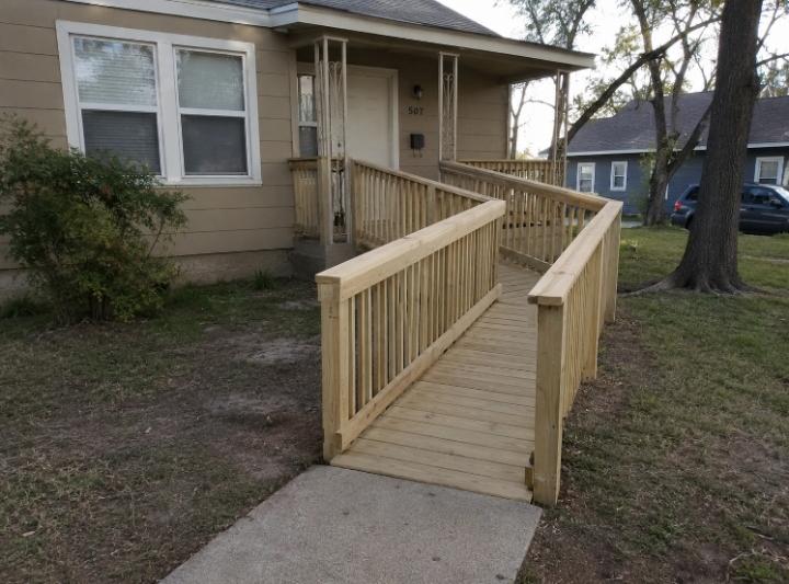 A newly installed wooden wheelchair ramp at the front door of a modest Texas home — natural light, residential setting