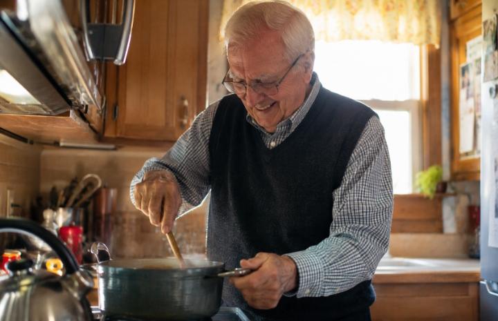 An elderly man with mild hand tremors smiling while stirring a pot in a warm, sunlit kitchen — candid, not posed, no clinical aesthetic