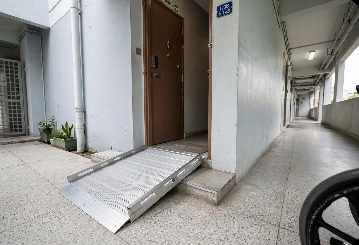 Photo of a small aluminum wheelchair ramp installed over a low step at an HDB flat entrance, daylight, wheelchair wheel partially in frame, clean and practical looking, no people